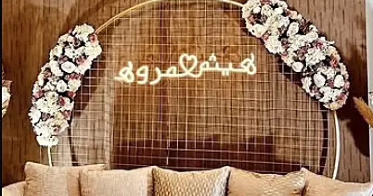 Arabic couple names in warm white neon on a floral wedding backdrop